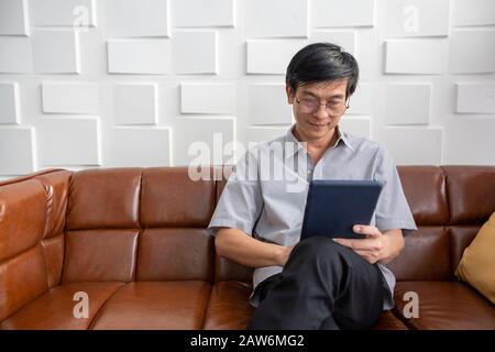 Senior Asian man playing tablet on sofa in living room at home  Portrait of Asian elderly man is Relaxing and Happiness With playing tablet Stock Photo