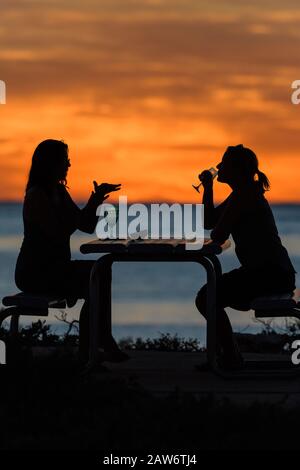 A bright orange sunset silhouetting two ecotourists enjoying a happy hour drink over the Indian Ocean at Osprey Bay, Cape Range National Park in WA. Stock Photo