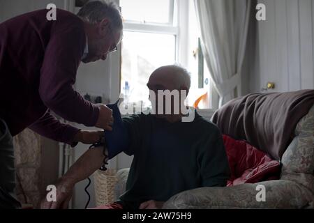 GP Dr David Binnie checking the blood pressure of patient Walter Williams at his home at Kiloran Bay on the the Inner Hebridean island of Colonsay on Scotland's west coast.  The island is in the council area of Argyll and Bute and has an area of 4,074 hectares (15.7 sq mi). Aligned on a south-west to north-east axis, it measures 8 miles (13 km) in length and reaches 3 miles (4.8 km) at its widest point, in 2019 it had a permanent population of 136 adults and children. Stock Photo