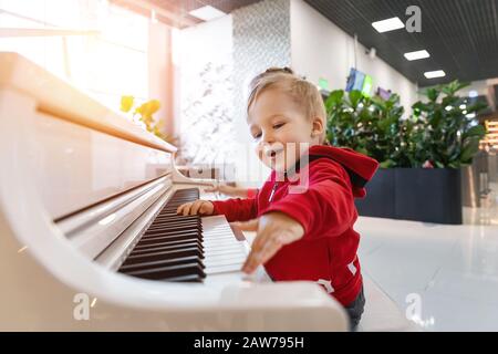 Adorable cute caucasian little toddler boy having fun playing big white piano in mall indoor. Funny happy small child enjoy singing and making loud