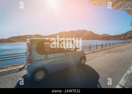 Eco car driving on road with lake and mountain landscape scenery. Motion blur showing car movement. Concept of road trip travel, rental cars, green en Stock Photo