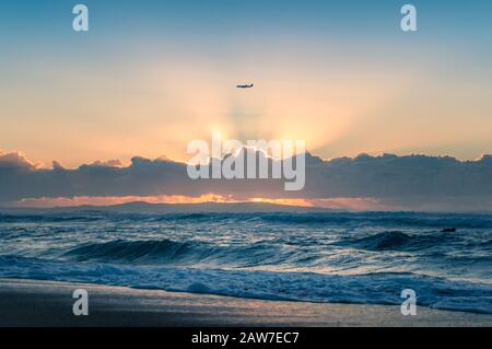 Silhouette of air plane with sun shining through clouds forming god rays and blue ocean beneath Stock Photo