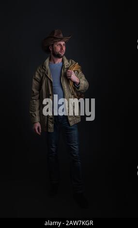 Cowboy in a wide-brimmed hat and jacket with a lasso on his shoulder posing on a dark background Stock Photo