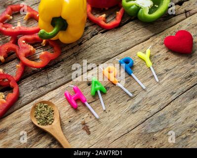 flat lay of cooking utensils with herbs , colorful bell pepper and word happy from candles over wooden background. top view with copy space. enjoy eat Stock Photo