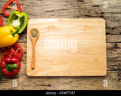 flat lay of empty blank wooden chopping board with wooden spoon and utensils with herbs and colorful bell pepper over wooden background. top view with Stock Photo