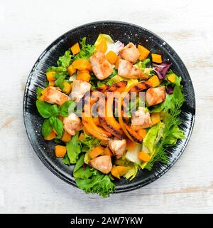 Pumpkin salad with chicken meat and vegetables in a black plate on a white wooden background. Top view. Free space for your text. Stock Photo