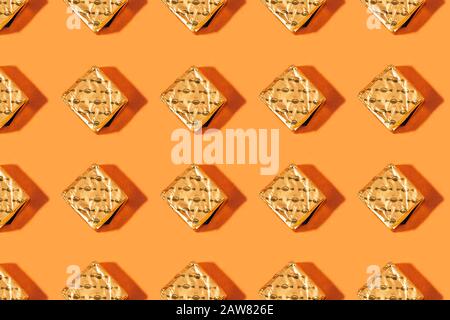 Chocolates pieces wrappers golden, on orange background, repeating pattern with hard shadows.