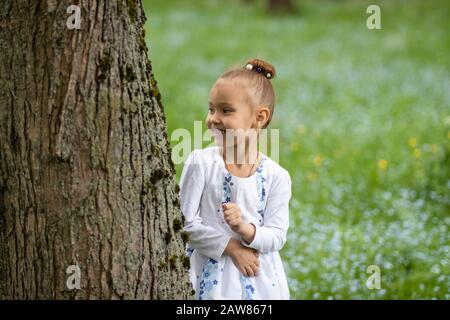 portrait of a cute little girl in a light dress on a background of an old tree.girl holding a small bouquet of forget-me-not flowers and smiling Stock Photo