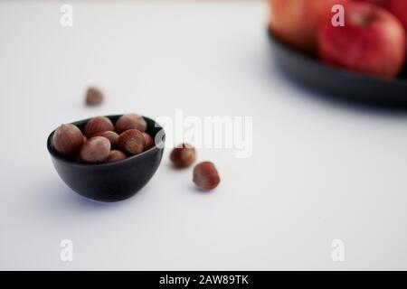 Hazelnuts in a dark colored bowl on a white background with red apples in the background Stock Photo