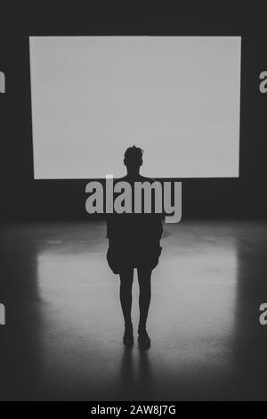 dramatic black and white image of a Silhouette of a female human figure standing with back to camera with an illuminated screen in the background