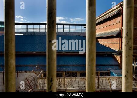 selective focus of view through iron bars to prison walls lined with barbed wire and rooftop security hall, during a sunny day Stock Photo
