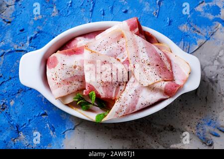 high angle view of some slices of raw bacon in a white ceramic bowl on a gray and blue textured surface Stock Photo