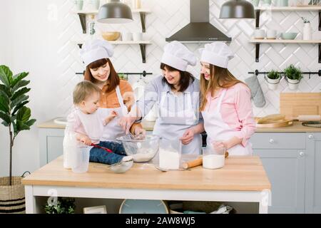 three generations of women in white aprons are playing and laughing while kneading the dough in the kitchen, mix flour with eggs and prepare dough in Stock Photo