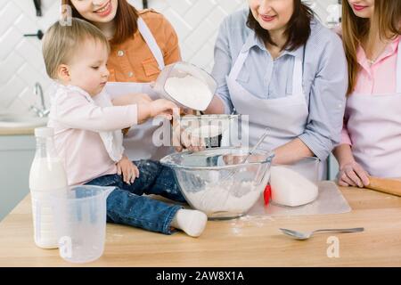 Adorable baby with mother, aunt and grandmother making dough with flour and egg and sugar together at home. Women in white aprons and chef hats sprink Stock Photo