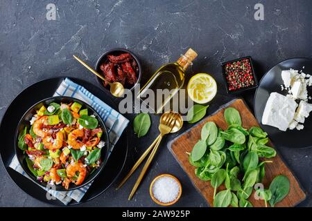 overhead view of shrimp avocado spinach salad with sun-dried tomatoes, crumbled cheese, red onion in a black bowl on a concrete table with ingredients Stock Photo