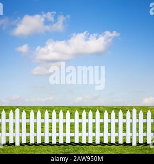 A white picket fence in front of a beautiful green field and blue sky in summer Stock Photo