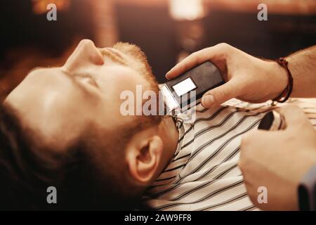 Close up Man is shaved in barbershop with electric razor. Hands of hairdresser frame holding tool Stock Photo