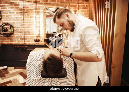 Barber man shaves his beard and cuts hair to client in barbershop. Stock Photo