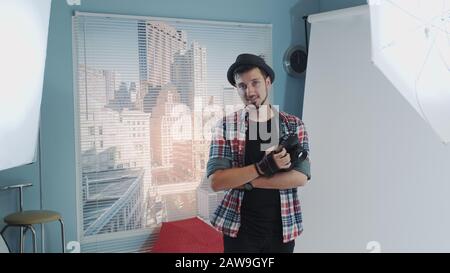 Handsome young photographer in hat holding camera and smiling in modern studio photo shoot. There are skyscrapers in the background. Stock Photo