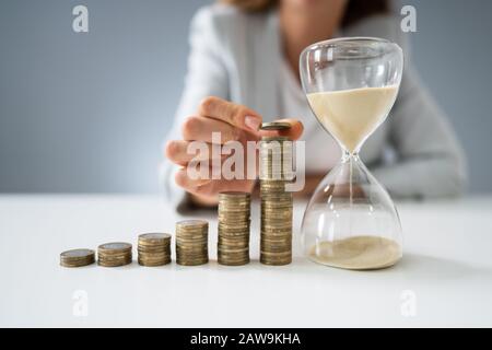 Close-up Of Businesswoman Holding Hourglass Near Stack Of Coins Stock Photo