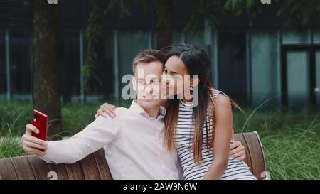 Romantic multiethnic couple sitting on bench in park and taking selfie. They smiling to the camera. Stock Photo
