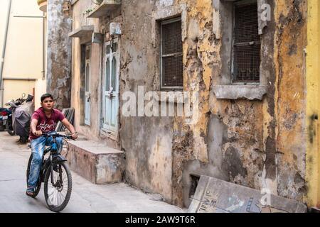 Diu, India - December 2018: A boy rides a bicycle down a narrow lane lined with faded, crumbling walls and windows of old houses. Stock Photo