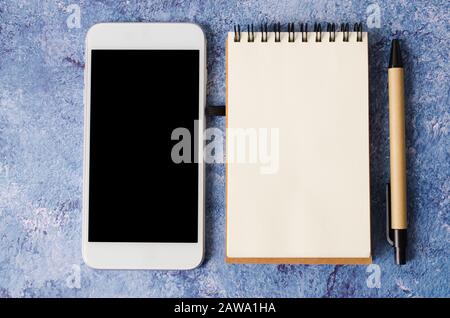 Empty notebook and smartphone on blue desk. Empty paper in notepad. Flat lay with copy space. Mock up. Stock Photo