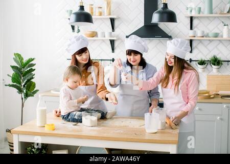 Happy women in white aprons baking together, Cutting out shapes from sugar cookie dough with cookie cutters. Little baby girl helps to make cookies to Stock Photo