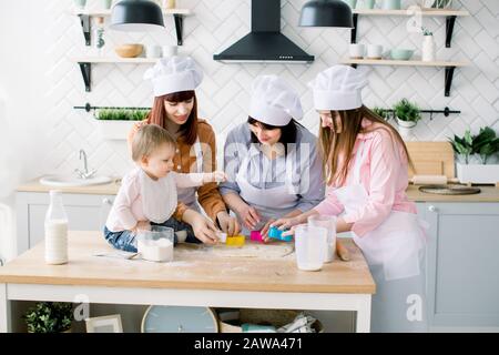 Happy women in white aprons baking together, Cutting out shapes from sugar cookie dough with cookie cutters. Little baby girl helps to make cookies to Stock Photo