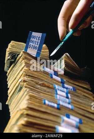 A surgeons hand holding a scalpel cutting through a stack of hundred dollar bills illustrating the high cost of medical care Stock Photo