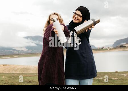 Lifestyle portrait of two best friends, smiling and drinking coffee together. Outdoor photo of two young women, one with hijab, enjoying each other co Stock Photo