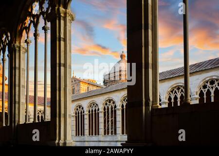 The Duomo Pisa Cathedral dome is visible under a colorful sunset from inside stone arched windows in the Tuscan city of Pisa, Italy.