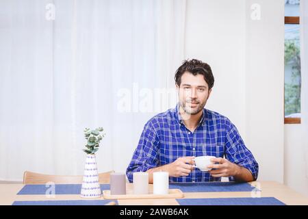gentle handsome man sitting in the kitchen table to drink some hot beverage or coffee with white space for text. Stock Photo