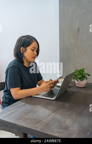 Asian female freelancer using smartphone and laptop at coffee shop Stock Photo