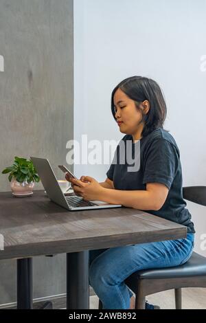 Asian female freelancer using smartphone while working on laptop Stock Photo