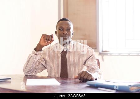 Sitting African Businessman working on documents on desk in front of office background Stock Photo