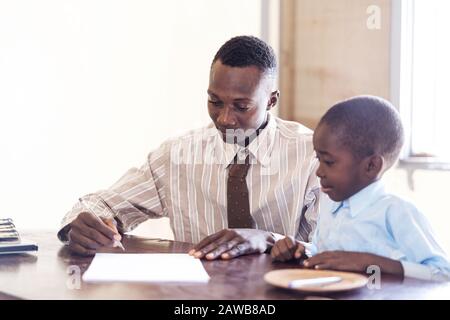 Cheerful young African man in shirt with his son while sitting against an office background Stock Photo