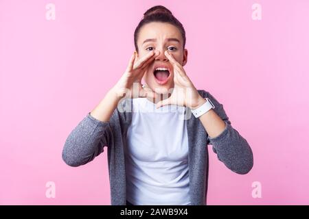 Attention! Portrait of brunette teenage girl with bun hairstyle in casual clothes holding hands near wide open mouth, loudly announcing ad, looking wo Stock Photo