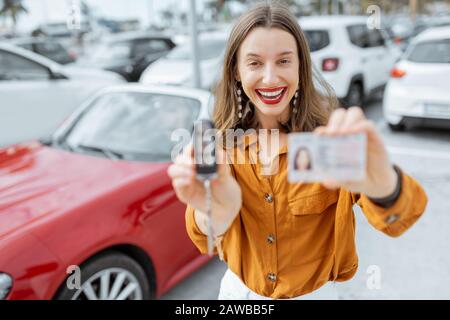 Portrait of a happy woman showing driver's license and keys standing near the red cabriolet at the car parking. Concept of a happy buying or renting a car Stock Photo