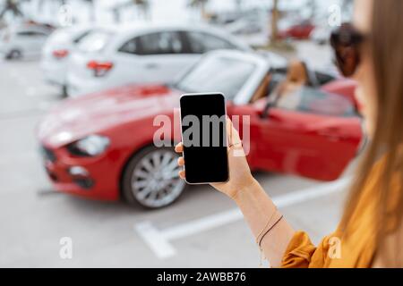 Woman holding a smartphone with empty screen at the car parking with red sports vehicle on the background, close-up view Stock Photo
