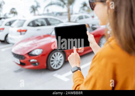 Woman controlling car with a digital tablet while standing near the vehicle outdoors, close-up view on the tablet with black screen Stock Photo