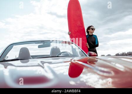 Portrait of a young woman surfer in swimsuit standing with surfboard behind her red car on the rocky coast Stock Photo