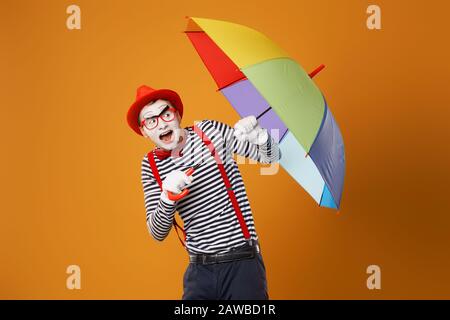 Surprised mime with multi-colored umbrella isolated on blank orange background in studio Stock Photo