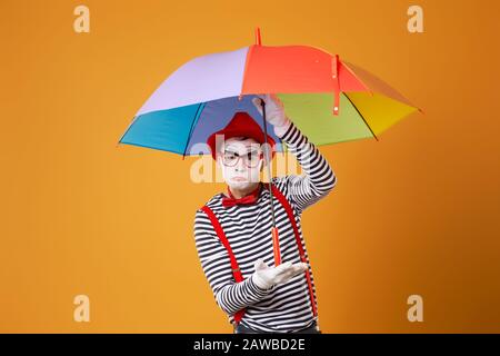 Serious mime with multi-colored umbrella on empty orange background in studio Stock Photo