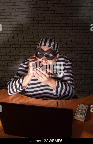 Comic character prisoner telephone con man sitting at a laptop in a striped robe and a hat in a black mask and cheating on the phone Stock Photo