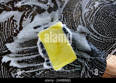 Hand in a black glove washes the glass with a foam sponge with foam on a dark background. Stock Photo