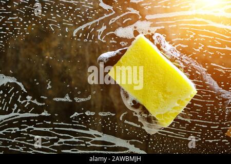 Hand in a black glove washes the glass with a foam sponge with foam on a dark background with light. Stock Photo