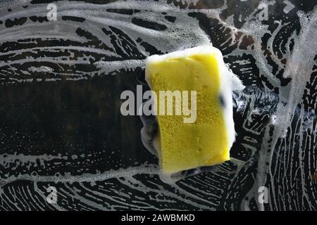 Hand in a black glove washes the glass with a foam sponge with foam on a dark background. Stock Photo
