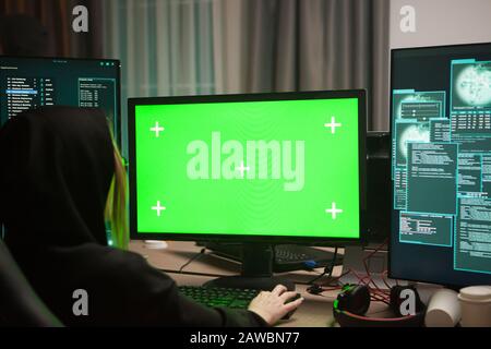 Wanted female hacker in front of computer with green screen. Dark room. Stock Photo