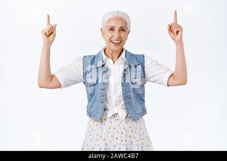Happy, cheerful charming grandmother, senior lady smiling and laughing carefree, raising hands, pointing fingers up, showing advertisment, promote Stock Photo